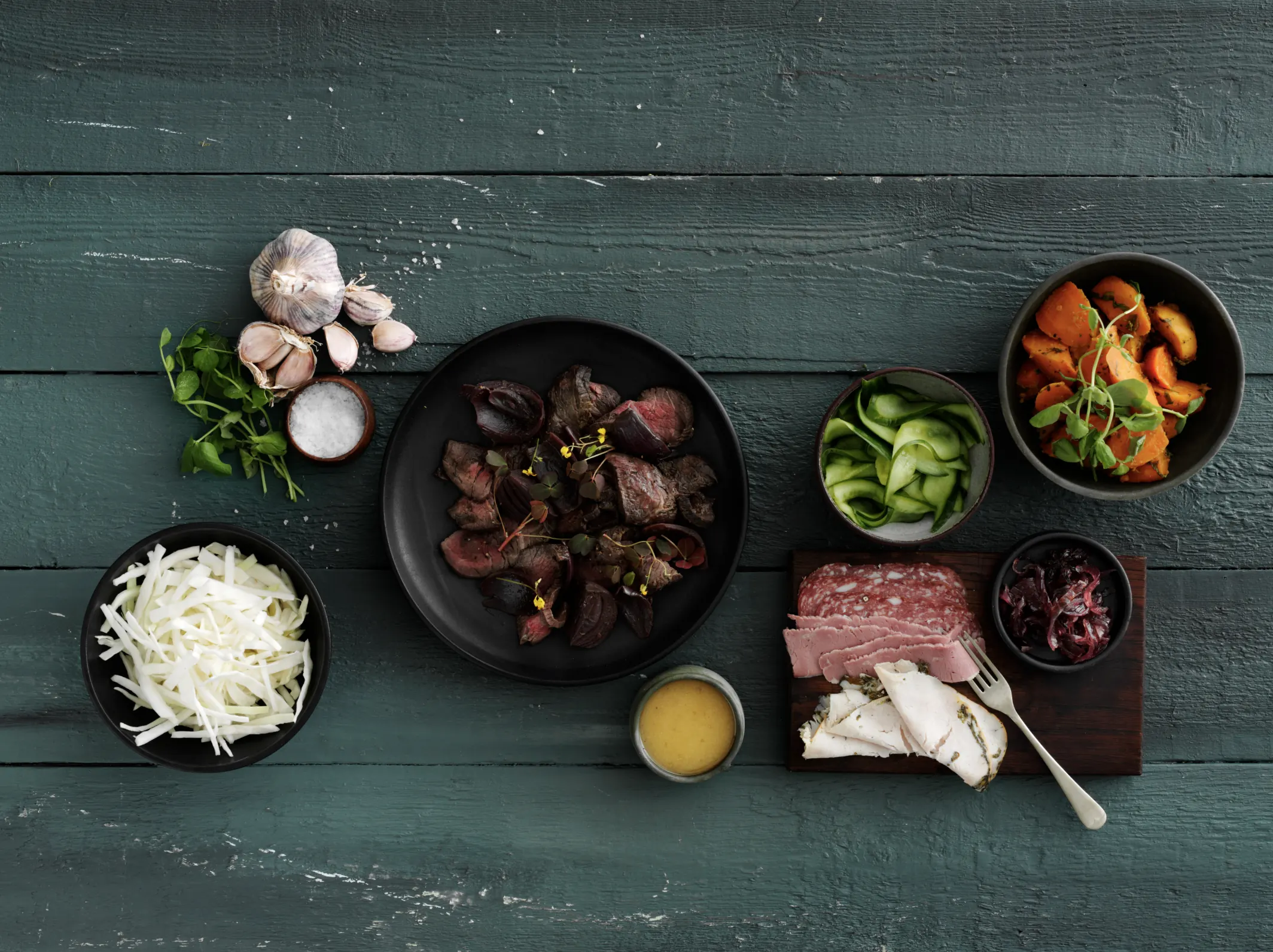 Frokostordning fra Frokostfirmaet: oksekød, pålæg, salater og tilbehør anrettet på grønt træbord